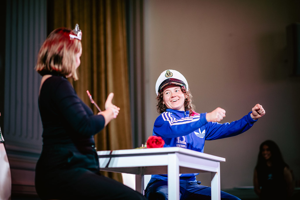 En person med kort grått hår och glasögon sitter i ett upplyst rum och talar i en mikrofon. Hen bär en färgstark, geometriskt mönstrad tröja i rött, orange och lila. I förgrunden syns siluetter av publikens huvuden, delvis suddiga. I bakgrunden står två personer som tolkar i teckenspråk, klädda i mörka kläder. Rummet har höga väggar och en varm belysning som skapar en högtidlig seminarie- eller panelsamtalsatmosfär.