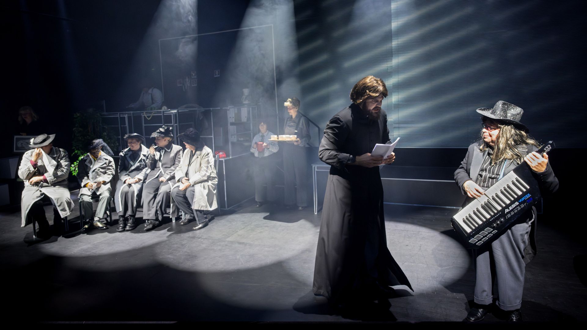En teaterscen med dramatisk punktbelysning visar flera skådespelare i en absurd, humoristisk situation. Till vänster sitter sex personer i grå trenchcoats och hattar på rad, som om de väntade på förhör eller instruktioner. Bakom dem arbetar andra skådespelare vid skrivbord fyllda med rekvisita, bland annat en röd telefon och en bricka med koppar. I förgrunden står en man klädd i lång svart dräkt och läser högt ur ett manus, medan en skådespelare till höger, klädd i glittrig hatt och lång peruk, spelar på ett keytar-liknande elektroniskt instrument. Scenen badar i dimma och ljusstrålar, vilket förstärker känslan av en stiliserad och lekfull kriminal- eller noirparodi.