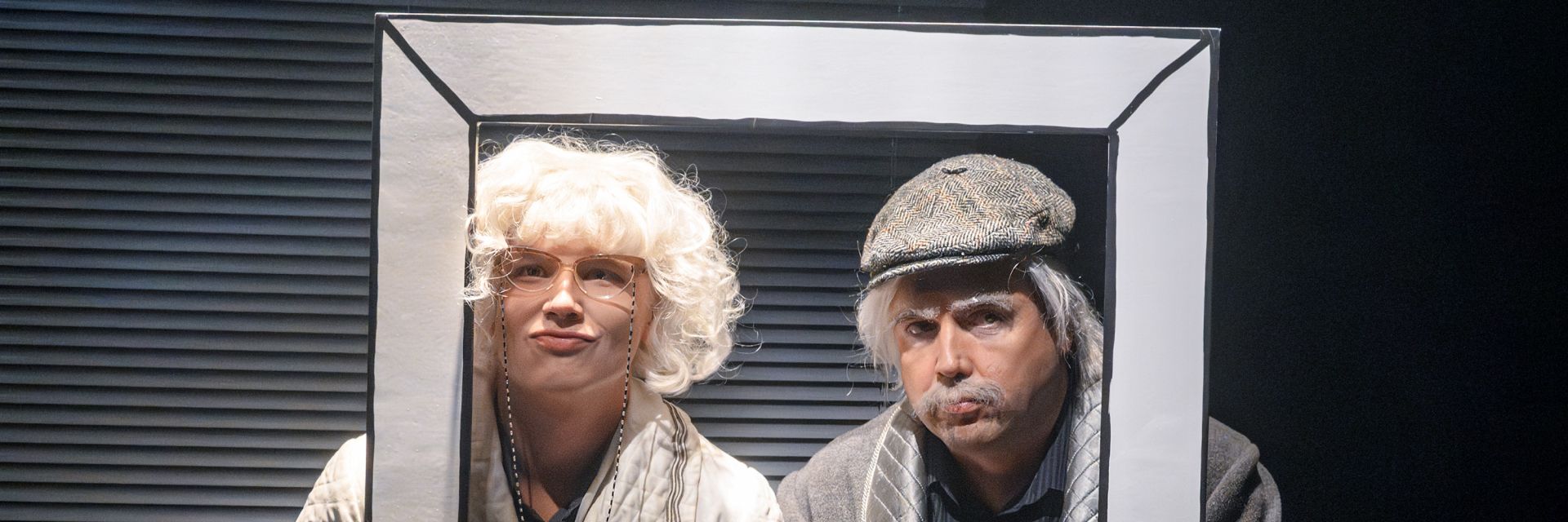 Två skådespelare står tätt intill varandra och tittar ut genom en rektangulär, målad pappersram som om de poserar för ett porträtt. Personen till vänster har lockig vit peruk, stora glasögon med kedja och gör en överdriven pussmun. Personen till höger bär keps i tweedmönster, grå peruk och buskiga vita ögonbryn samt mustasch, och ser skeptiskt åt sidan. Bakgrunden är mörk med horisontella skuggor, vilket framhäver de humoristiska mimikerna i förgrunden.