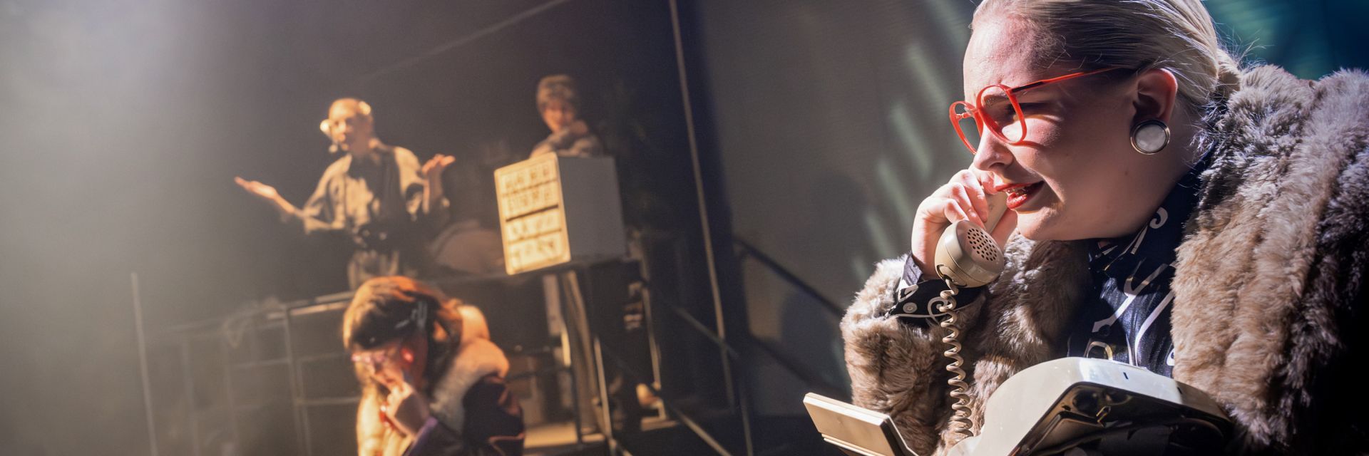 En skådespelare i förgrunden sitter i pälsjacka och stora röda glasögon, och pratar intensivt i en vit bordstelefon medan scenljuset faller starkt över ansiktet. I mitten av bilden sitter en annan person, också i pälsdetaljer, i en koncentrerad telefonkonversation. Längre bak på scenen står två skådespelare i dimmigt ljus; den ena gör en uppgiven gest med armarna, medan den andra står vid en apparat som liknar en gammaldags kontorsmaskin. Scenen har en hektisk och kaotisk atmosfär som påminner om ett kontorslandskap i kris.