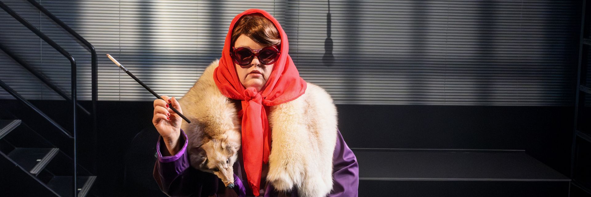 En skådespelare står i scenljus iförd stora röda solglasögon, en röd huvudscarf knuten under hakan och en lång ljus pälsboa med ett rävdjurshuvud framtill. Hen bär en lila rock och håller upp en lång cigarettmunstycke i höger hand med en dramatisk, nästan aristokratisk gest. Bakgrunden består av en mörk trappa och en vägg med horisontella skuggade linjer som skapar en industriell och film noir-liknande stämning.
